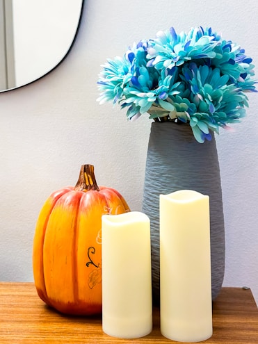 A wooden surface adorned with decorative items including a rustic orange pumpkin, two cream-colored pillar candles, and a bouquet of blue-hued flowers in a textured gray vase. A round mirror is partially visible in the background.