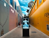 A walkway between two high-fashion branded walls with large advertisements for Tiffany & Co. and Fendi. A digital directory board displaying a fashion advertisement for Dior is placed in the center. A person carrying a shopping bag walks along the path. The sky above is bright with scattered clouds.