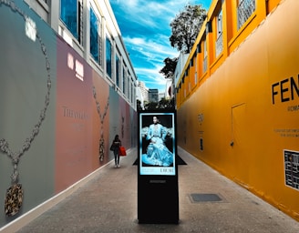A walkway between two high-fashion branded walls with large advertisements for Tiffany & Co. and Fendi. A digital directory board displaying a fashion advertisement for Dior is placed in the center. A person carrying a shopping bag walks along the path. The sky above is bright with scattered clouds.