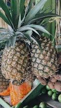 A close-up view of two ripe pineapples with green spiky leaves. The setting includes various tropical fruits such as avocados arranged below. Behind, there are jars filled with pickled vegetables.