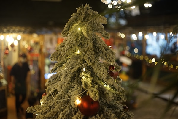 A close-up of a frosted artificial Christmas tree adorned with small lights and red ornaments. The background is blurred, showcasing warm indoor lighting and hints of festive decorations, creating a cozy and seasonal ambiance.