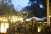 A tranquil outdoor scene with a series of wooden birdhouses arranged on a wooden platform. In the background, soft bokeh lights create a warm ambiance, and blurred foliage suggests a garden setting. An illuminated lamp on the left adds a cozy glow to the surroundings.