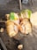 A freshly cut palm fruit is opened to reveal its fibrous interior and seed segments. The outer skin displays shades of green and brown, while the interior is a lighter beige with cluster-like white seeds. The fruit rests on a rough wooden surface with other pieces scattered around, suggesting a natural and rustic setting.