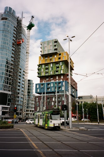 A modern cityscape featuring a colorful high-rise building with a unique block design. A tram is visible on the street intersection, surrounded by other skyscrapers and construction cranes. The scene captures urban architecture and transportation.