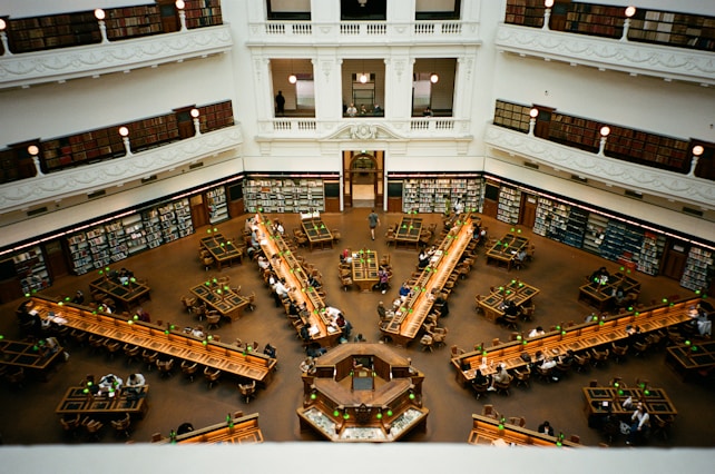 A spacious library with high ceilings and multiple levels of bookshelves. The central reading area features long wooden tables and chairs, occupied by people studying or reading. The architecture is classical, with ornate details and large, arched windows allowing natural light to filter in.