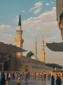 A majestic mosque with multiple minarets and a prominent green dome is set against a backdrop of a partly cloudy sky. The plaza is busy with people walking, dressed in a variety of traditional and modern clothing. Large umbrella-like structures provide shade over the open courtyard area.
