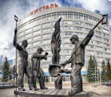 A large metal sculpture depicting three workers is prominently placed in front of a multi-story glass and concrete building with the word 'Ижсталь' visible on the top. The workers are shown as statues with two holding long tools and one standing with crossed arms, symbolizing industrial labor. Evergreen trees are planted in front of the building, adding a natural element to the urban landscape. The sky above is partly cloudy, casting soft light on the scene.