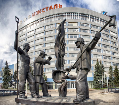A large metal sculpture depicting three workers is prominently placed in front of a multi-story glass and concrete building with the word 'Ижсталь' visible on the top. The workers are shown as statues with two holding long tools and one standing with crossed arms, symbolizing industrial labor. Evergreen trees are planted in front of the building, adding a natural element to the urban landscape. The sky above is partly cloudy, casting soft light on the scene.