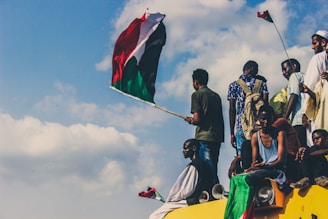 A group of people are gathered on top of a vehicle. One person is prominently waving a large flag with red, white, and green colors. The sky is bright with scattered clouds, and some individuals are holding smaller flags. The scene conveys a sense of unity and collective action.