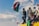 A group of people are gathered on top of a vehicle. One person is prominently waving a large flag with red, white, and green colors. The sky is bright with scattered clouds, and some individuals are holding smaller flags. The scene conveys a sense of unity and collective action.