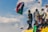 A group of people are gathered on top of a vehicle. One person is prominently waving a large flag with red, white, and green colors. The sky is bright with scattered clouds, and some individuals are holding smaller flags. The scene conveys a sense of unity and collective action.