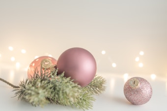 Pink and gold Christmas ornaments rest on a surface next to a sprig of evergreen. Soft, blurred lights in the background create a warm, festive atmosphere.