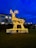 A large, illuminated reindeer sculpture made of geometric shapes is situated on a grassy area, surrounded by a white picket fence. The sculpture is aglow with warm yellow lights against a deep blue evening sky. In the background, a building with lit windows and some trees can be seen.