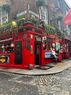 A vibrant red pub situated on a corner street, adorned with various outdoor signs and plaques. The building facade features hanging plants under the windows and a variety of advertising signs for drinks. A man in casual attire is sitting outdoors near the entrance. The cobblestone street adds a rustic charm to the scene.