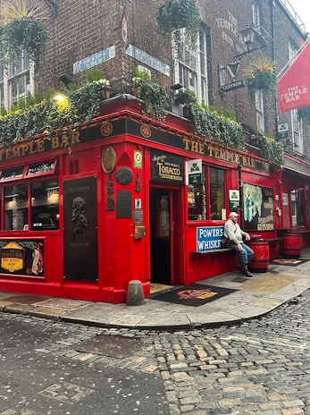 A vibrant red pub situated on a corner street, adorned with various outdoor signs and plaques. The building facade features hanging plants under the windows and a variety of advertising signs for drinks. A man in casual attire is sitting outdoors near the entrance. The cobblestone street adds a rustic charm to the scene.