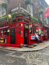 A vibrant red pub situated on a corner street, adorned with various outdoor signs and plaques. The building facade features hanging plants under the windows and a variety of advertising signs for drinks. A man in casual attire is sitting outdoors near the entrance. The cobblestone street adds a rustic charm to the scene.