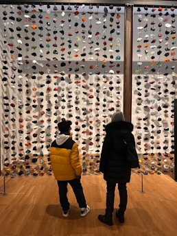 Two people stand in front of a large hanging installation composed of numerous colorful stones or glass pieces strung vertically. They are inside a well-lit exhibition space with wooden flooring, and their backs are to the viewer as they observe the art piece.