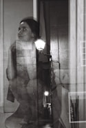 A black and white double exposure photograph featuring a woman with an introspective expression blended with an alley scene illuminated by streetlights. Her gaze is directed upward with her chin resting on her hand, while the textures of brick walls and pavement add depth to the composition.