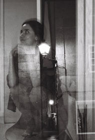 A black and white double exposure photograph featuring a woman with an introspective expression blended with an alley scene illuminated by streetlights. Her gaze is directed upward with her chin resting on her hand, while the textures of brick walls and pavement add depth to the composition.