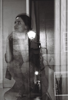 A black and white double exposure photograph featuring a woman with an introspective expression blended with an alley scene illuminated by streetlights. Her gaze is directed upward with her chin resting on her hand, while the textures of brick walls and pavement add depth to the composition.