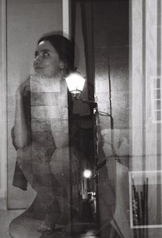 A black and white double exposure photograph featuring a woman with an introspective expression blended with an alley scene illuminated by streetlights. Her gaze is directed upward with her chin resting on her hand, while the textures of brick walls and pavement add depth to the composition.