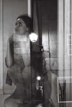A black and white double exposure photograph featuring a woman with an introspective expression blended with an alley scene illuminated by streetlights. Her gaze is directed upward with her chin resting on her hand, while the textures of brick walls and pavement add depth to the composition.