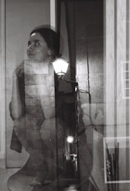 A black and white double exposure photograph featuring a woman with an introspective expression blended with an alley scene illuminated by streetlights. Her gaze is directed upward with her chin resting on her hand, while the textures of brick walls and pavement add depth to the composition.