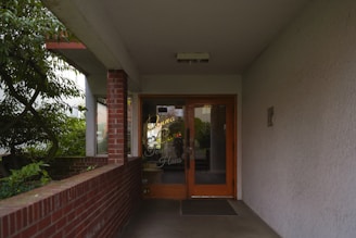 An entrance to a building featuring double glass doors with wooden frames. The doors have gold letters spelling out a name. The surrounding area includes a brick wall on one side and a smooth wall on the other, with some green foliage visible directly outside the open corridor.
