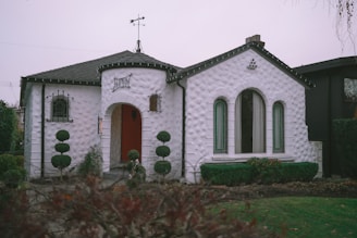 A quaint white stucco house with a unique rounded design and an inviting arched entrance. The architecture features textured walls and narrow, tall windows with green trim. A small weather vane sits atop the roof, and neatly trimmed topiary bushes flank the entrance, adding charm to the front garden.