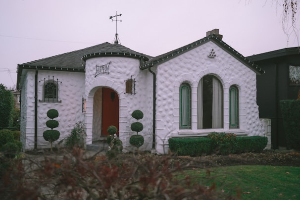 A quaint white stucco house with a unique rounded design and an inviting arched entrance. The architecture features textured walls and narrow, tall windows with green trim. A small weather vane sits atop the roof, and neatly trimmed topiary bushes flank the entrance, adding charm to the front garden.