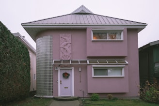 A two-story house with a unique architectural design, featuring a rounded corner with glass blocks and a light pink exterior. The house has a metal roof and two rectangular windows on the upper floor. There is a decorative panel with an abstract design beside the door, which is adorned with a wreath.