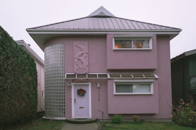 A two-story house with a unique architectural design, featuring a rounded corner with glass blocks and a light pink exterior. The house has a metal roof and two rectangular windows on the upper floor. There is a decorative panel with an abstract design beside the door, which is adorned with a wreath.