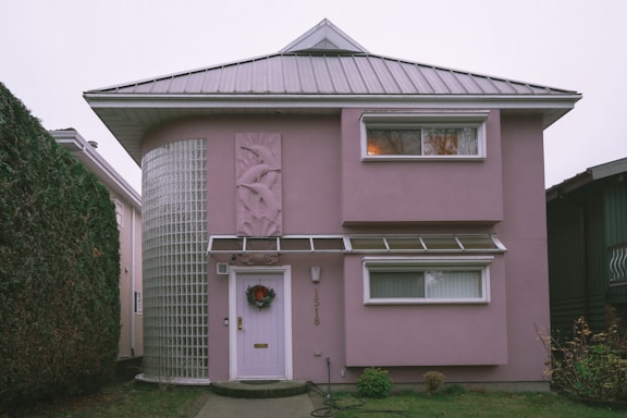 A two-story house with a unique architectural design, featuring a rounded corner with glass blocks and a light pink exterior. The house has a metal roof and two rectangular windows on the upper floor. There is a decorative panel with an abstract design beside the door, which is adorned with a wreath.