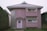 A two-story house with a unique architectural design, featuring a rounded corner with glass blocks and a light pink exterior. The house has a metal roof and two rectangular windows on the upper floor. There is a decorative panel with an abstract design beside the door, which is adorned with a wreath.