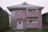 A two-story house with a unique architectural design, featuring a rounded corner with glass blocks and a light pink exterior. The house has a metal roof and two rectangular windows on the upper floor. There is a decorative panel with an abstract design beside the door, which is adorned with a wreath.
