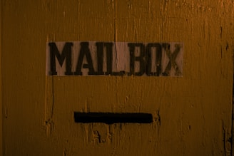 A wooden surface is painted in a rough, rustic style with a dark yellow or mustard color. The word 'MAILBOX' is stenciled in black letters on a white background. Below the text, there is a narrow horizontal slot designed for inserting mail.