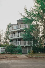 A multi-story apartment building with circular balconies, surrounded by lush green trees and shrubs. The entrance is gated, and the sidewalk in front is covered with fallen leaves.