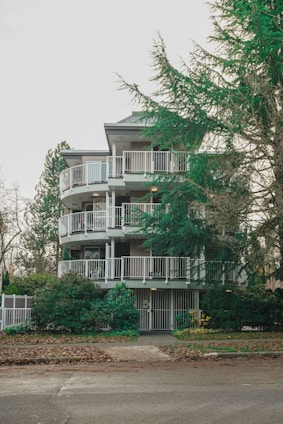 A multi-story apartment building with circular balconies, surrounded by lush green trees and shrubs. The entrance is gated, and the sidewalk in front is covered with fallen leaves.