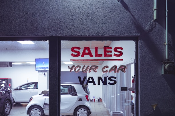 The interior of a car dealership is visible through a glass window that has bold lettering advertising sales of cars and vans. Several vehicles, including a white compact car, are arranged inside a well-lit showroom. A television screen mounted on the wall displays an image, while the room also contains various equipment and items.