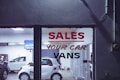 The interior of a car dealership is visible through a glass window that has bold lettering advertising sales of cars and vans. Several vehicles, including a white compact car, are arranged inside a well-lit showroom. A television screen mounted on the wall displays an image, while the room also contains various equipment and items.