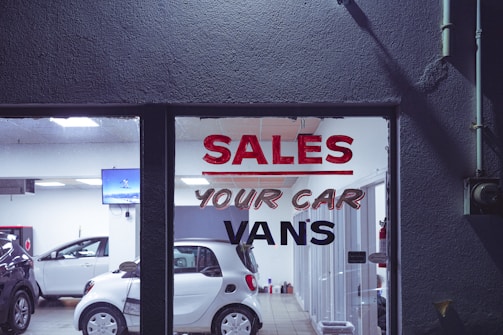 The interior of a car dealership is visible through a glass window that has bold lettering advertising sales of cars and vans. Several vehicles, including a white compact car, are arranged inside a well-lit showroom. A television screen mounted on the wall displays an image, while the room also contains various equipment and items.