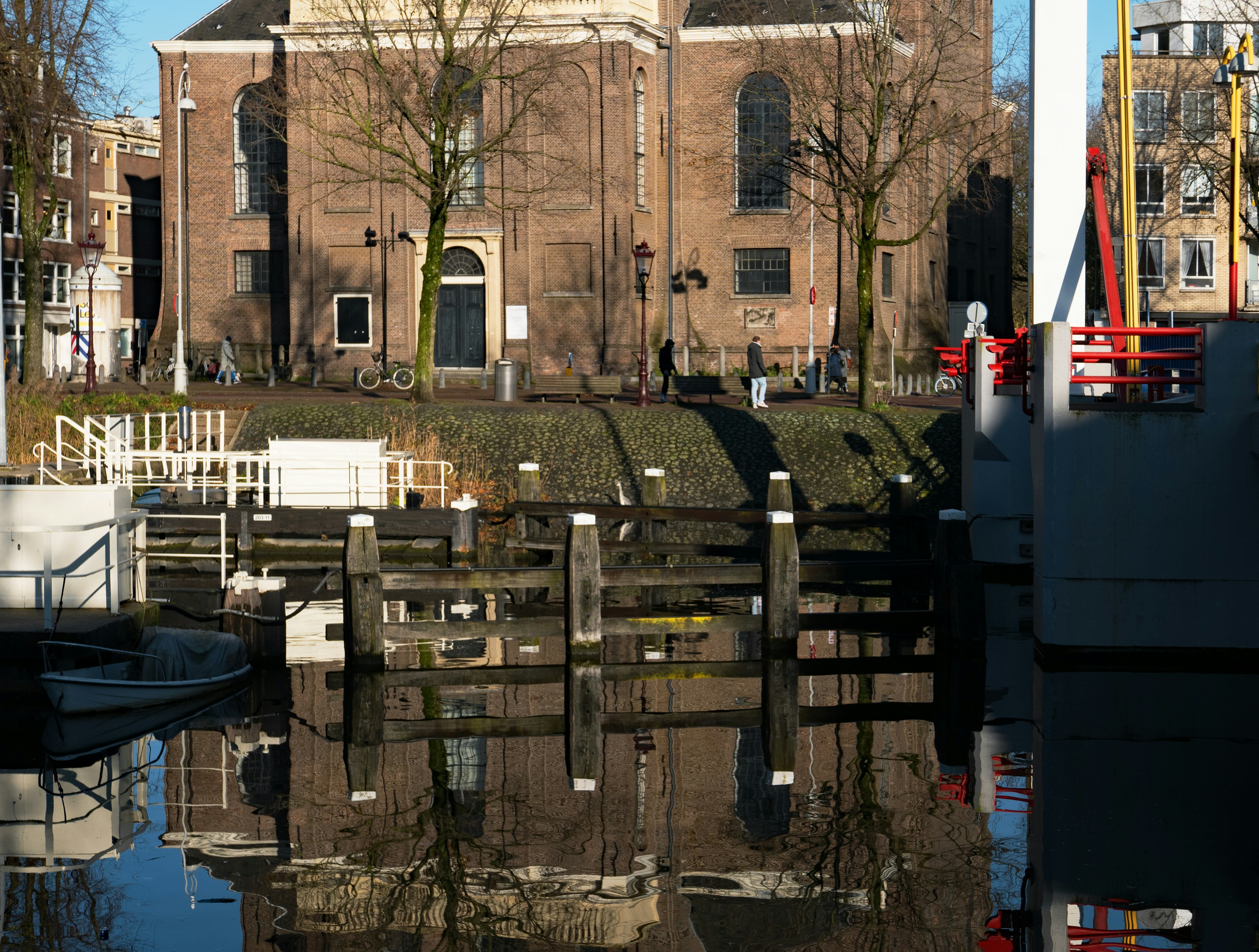 a building with a clock tower reflected in a body of water, Photo of the brick facade of the old church Oosterkerk in Amsterdam city. This is a historical church, because it is the first Amsterdam church built in a protestant architecture style. In front of the church is the reflecting canal water of the Nieuwe Vaart. Street photography of urban cities in The Netherlands by Fons Heijnsbroek, 2023 // Foto van Amsterdam, gratis download: Een foto in de zon van de oud bakstenen gevel van de historische Oosterkerk Het is herfst met veel zon aan de Wittenburgergracht; rechts de Nijlpaardenbrug - de sluis staat open. Nederland.