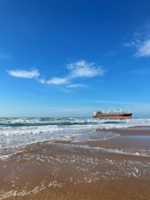 A large cargo ship sails on the horizon under a clear blue sky with a few scattered clouds. The ship is positioned over calm waves approaching a sandy beach, creating a peaceful seaside scene.