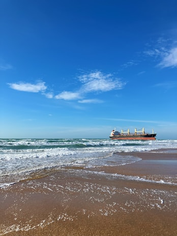 A large cargo ship sails on the horizon under a clear blue sky with a few scattered clouds. The ship is positioned over calm waves approaching a sandy beach, creating a peaceful seaside scene.