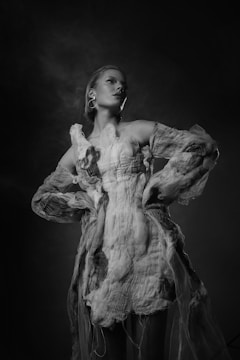 A figure stands confidently against a dark, smoky background. The person wears an abstract, textured garment that appears to be made from layers of fabric, giving the impression of an avant-garde fashion statement. The lighting highlights the contours of the face and the ethereal qualities of the outfit, creating a striking visual contrast.