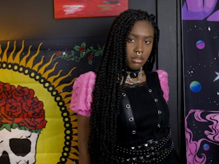 A person with long braided hair stands in front of a colorful backdrop. They are wearing a black outfit with a studded choker and layered necklaces. The background includes vibrant artwork, featuring a skull with red roses and planets against a cosmic theme.