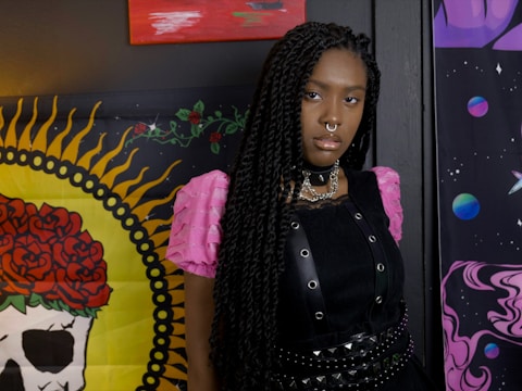 A person with long braided hair stands in front of a colorful backdrop. They are wearing a black outfit with a studded choker and layered necklaces. The background includes vibrant artwork, featuring a skull with red roses and planets against a cosmic theme.