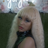 A person with long, wavy blonde hair is wearing a shiny black outfit. They have bold makeup with red lipstick and long eyelashes. Behind them is a decorative backdrop featuring roses and a sign with cursive white neon lettering.