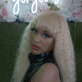 A person with long, wavy blonde hair is wearing a shiny black outfit. They have bold makeup with red lipstick and long eyelashes. Behind them is a decorative backdrop featuring roses and a sign with cursive white neon lettering.