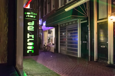 A brightly lit sign displaying the words 'coffee shop de kroon' is mounted on a building facade. The street appears to be deserted, with some dimly lit windows and a parked bicycle in the background. The scene is captured at night, with a cozy and quiet urban atmosphere.